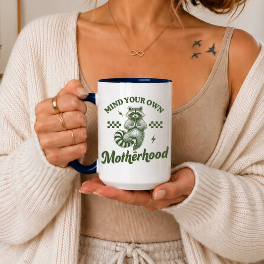 Woman holding a white mug with a raccoon illustration and text 'Mind Your Own Motherhood'