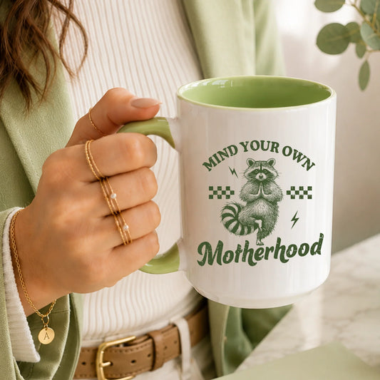 Hand holding a white and green mug with a raccoon and 'Mind Your Own Motherhood' text.
